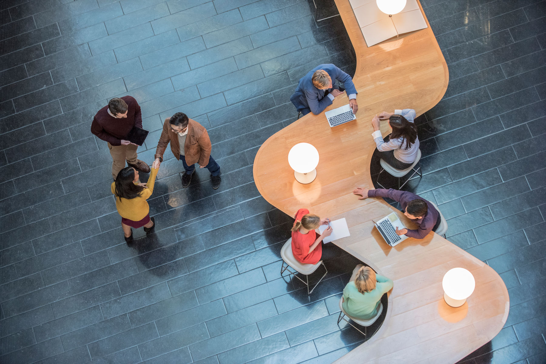 Aerial view of people working and meeting at a curved wooden desk with lamps. Two people are shaking hands whilst others sit and use laptops or write. The floor is dark tile.