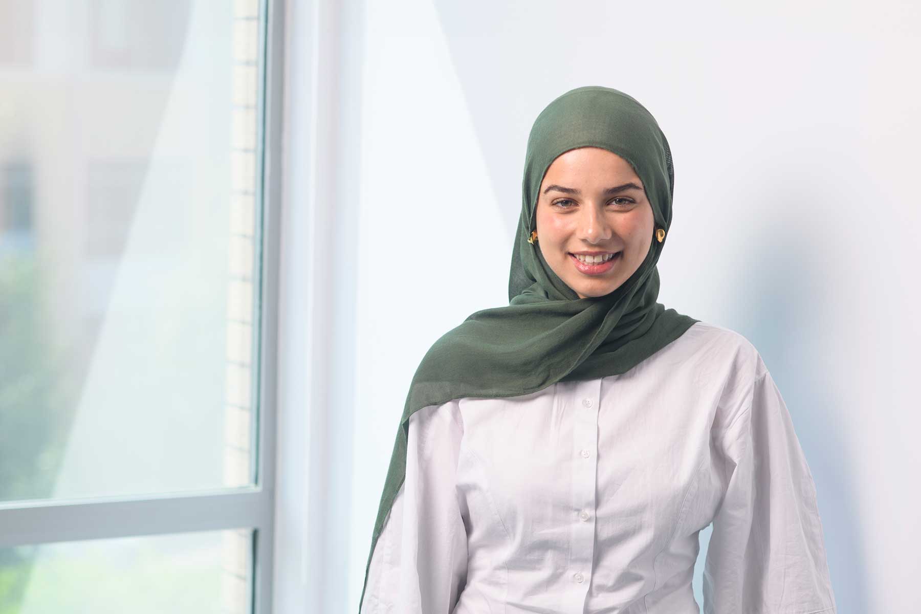 Hana Vorajee. A woman wearing a green hijab and white shirt smiles whilst standing indoors near a large window with soft natural light.