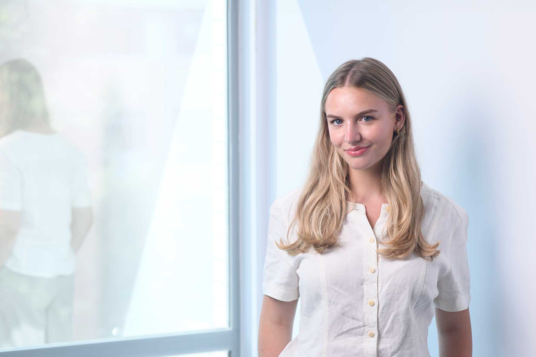 Imogen-Faye Earp. A young woman with long blonde hair and a white button-up shirt stands indoors by a window, smiling slightly.