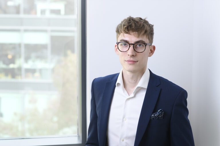 A young man with short brown hair and glasses, wearing a navy blue suit and white shirt, stands indoors near a window with blurred buildings outside. This is Josh Dunford.