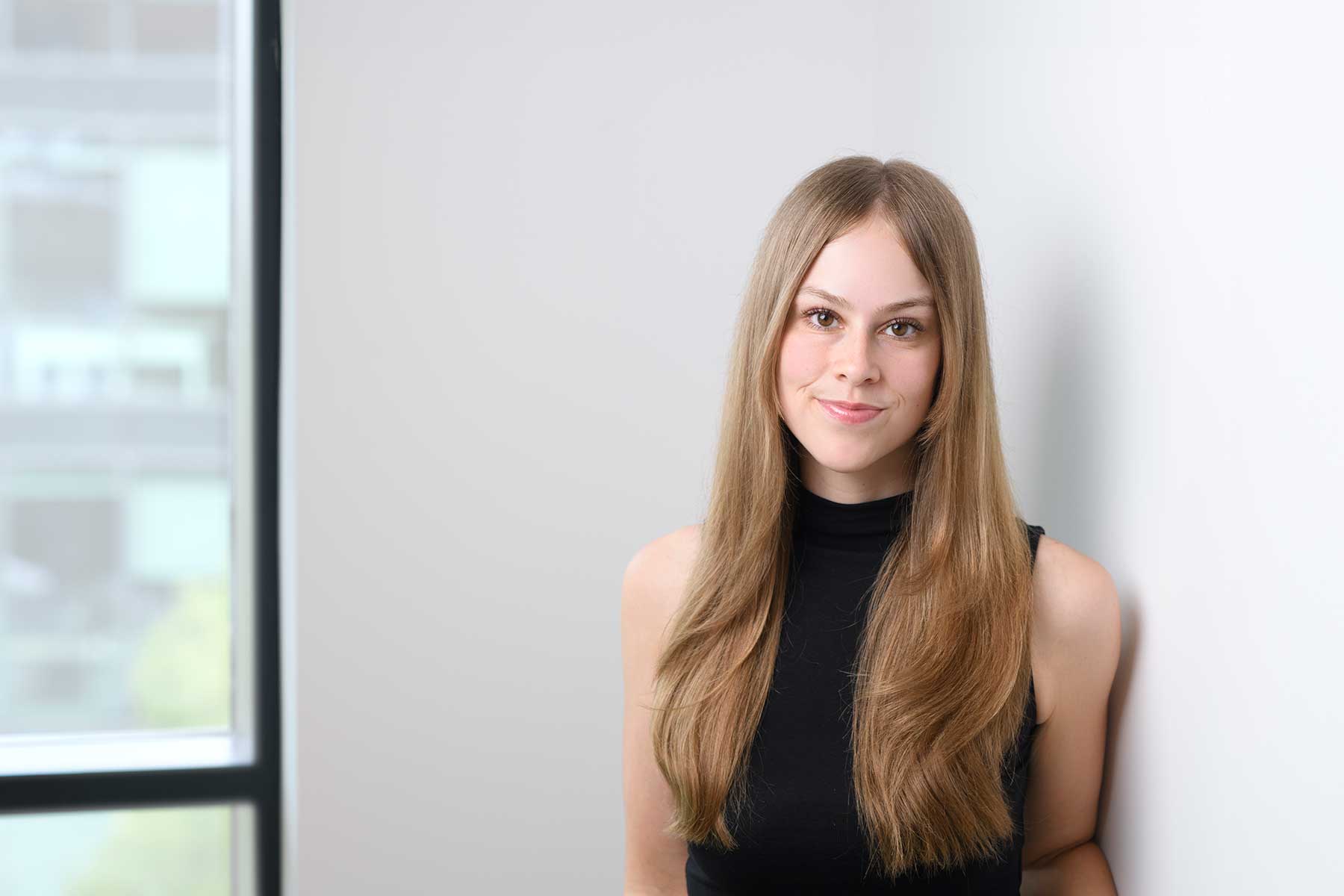 Lauren Tandy. Young woman with long straight hair, wearing a sleeveless black top, stands indoors against a white wall near a window, softly smiling at the camera.