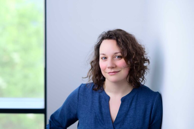 Sarah Pateman, A woman with curly brown hair wearing a blue top leans against a white wall, smiling softly. A window with a blurred green outdoor background is on the left.