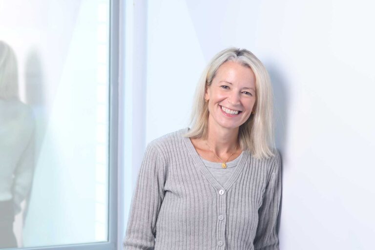 Selina Yelland, A smiling woman with straight blonde hair, wearing a grey ribbed cardigan, stands indoors leaning against a white wall near a window.