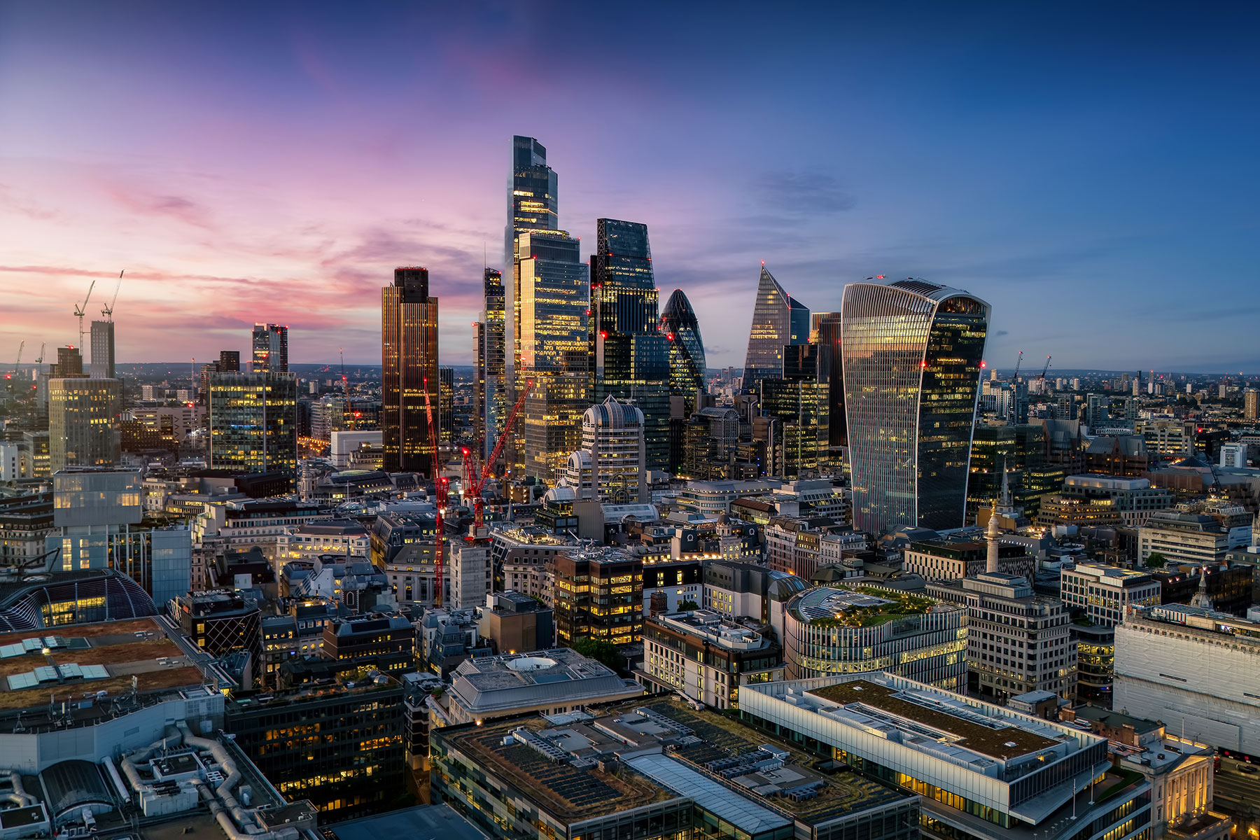 Aerial view of the London skyline at dusk, featuring prominent skyscrapers illuminated against a purple and blue sunset.