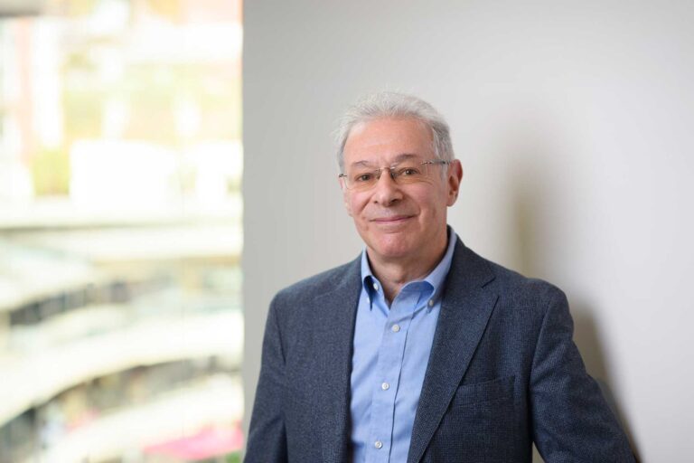 Gordon Nardell. An man with grey hair and glasses, wearing a blue shirt and blazer, stands indoors near a window, smiling softly at the camera.