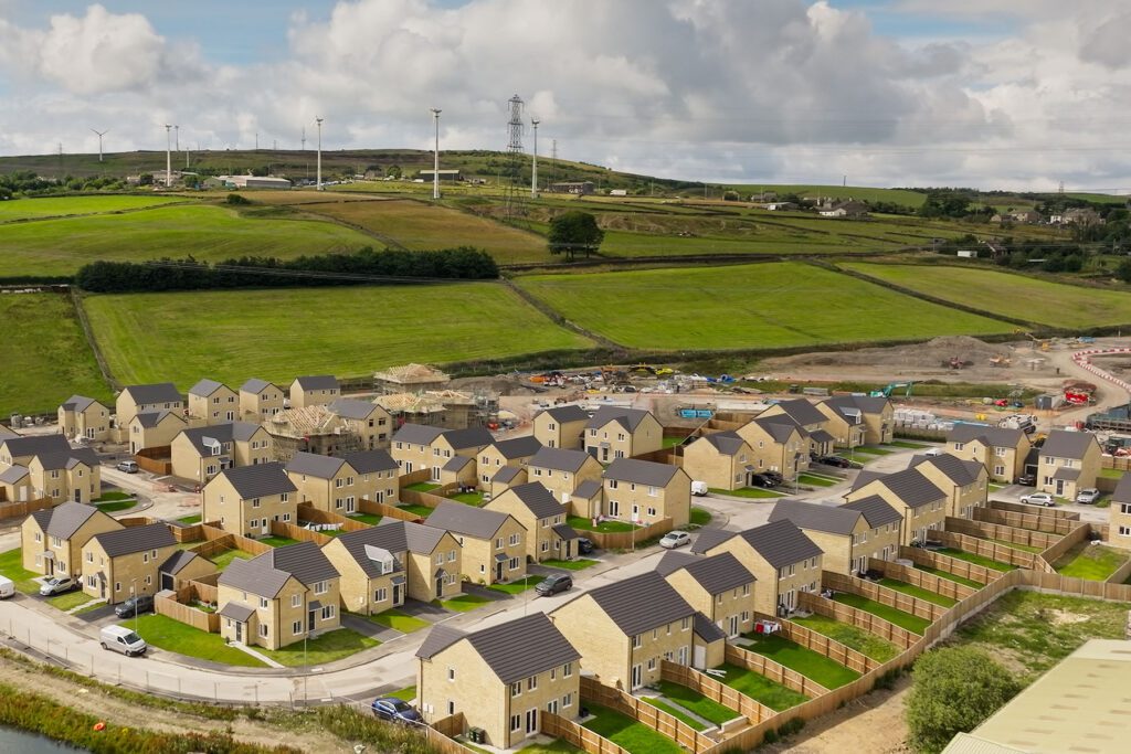 Aerial view of a new housing development with multiple tan-brick houses, fenced gardens, and construction activity in the background.