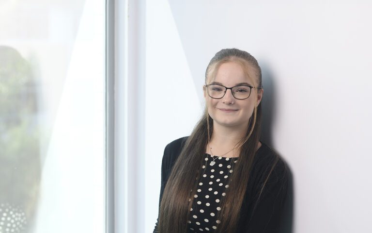 A woman with long brown hair, glasses, and a black and white spotty top stands by a large window, smiling softly at the camera.